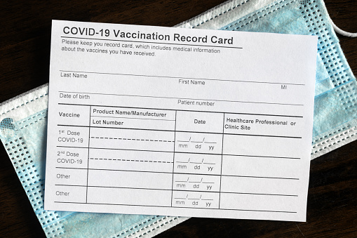 COVID-19 Vaccination Record Card on desk, coronavirus immunization certificate and surgical mask. Top view of paper medical form required for travel. Concept of corona virus vaccine, safety, tourism.