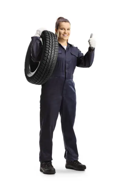 Full length portrait of a female mechanic worker carrying a tire and showing thums up isolated on white background