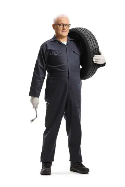 Full length portrait of a man auto mechanic carrying a tire and a lug wrench isolated on white background