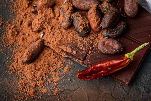 Dark chocolate, red chilli peppers, cocoa beans and powder on a dark stone background, top view, selective focus.