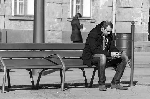 Timisoara, Romania - March 14, 2016: Man sitting on a bench on the street and listening to music from his phone. Real people. Black and white.