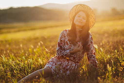 One beautiful young woman in dress with floral pattern and straw hat sitting and enjoying summer sunset over blooming meadow, feeling free and relaxed