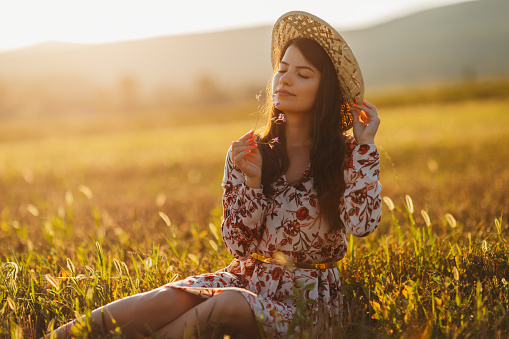 One beautiful young woman in dress with floral pattern and straw hat sitting and enjoying summer sunset over blooming meadow, feeling free and relaxed
