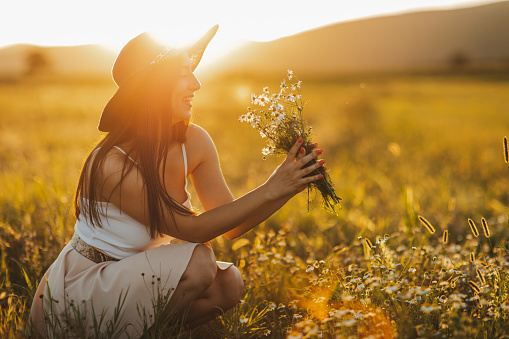 One beautiful young woman with sun hat crouching and smelling wildflower, enjoying summer sunset over blooming meadow, feeling free and relaxed, side view