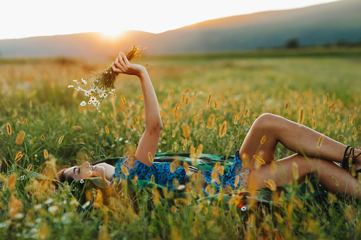 One beautiful young woman with bouquet of wildflowers, lying on picnic blanket and enjoying summer sunset over blooming meadow, feeling free, relaxed and happy in nature, side view