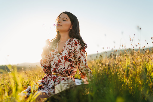 One beautiful young woman in dress with floral pattern sitting and enjoying summer sunset over blooming meadow, feeling free and relaxed