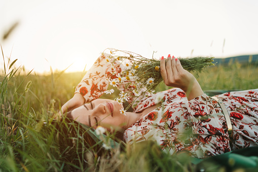 One beautiful young woman in dress with floral pattern lying, holding and smelling bouquet of wildflowers, enjoying summer sunset over blooming meadow, feeling free and relaxed, front view