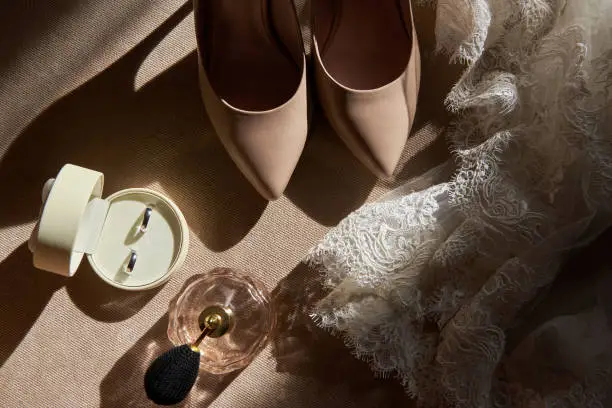 Close up of beige women shoes, two golden wedding rings in box, dress and perfume on chair, copy space. Wedding concept. Top view, flat lay
