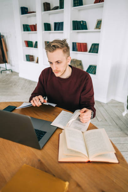 istock Young caucasian man college student studying with laptop distantly preparing for test exam writing essay doing homework at home or college library. 1310794608