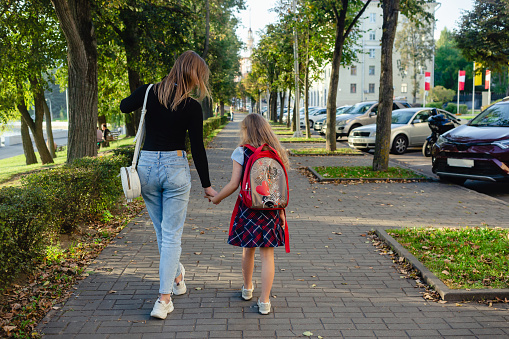 Adorable cute little girl feeling excited about going back from school with her mother by the hand. A pretty girl with a backpack walks on the street with her mother.