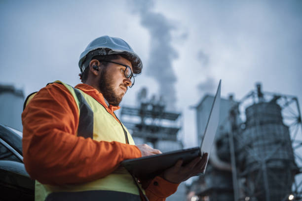 Serious handsome engineer using a laptop while working in the oil and gas industry. A shot of an young engineer wearing a helmet and using a laptop and hands free device during his night shirt in the oil rafinery. Engineering concept. chemical stock pictures, royalty-free photos & images