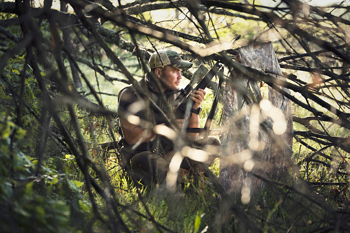 A shot of a young man with weapon hidding behind a tree in a forest.