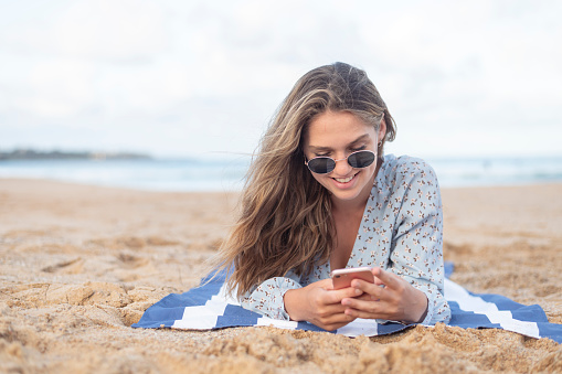 A beautiful and happy young adult woman uses her mobile phone to send a text or use a social media application while relaxing at the beach. The woman is lying on a beach towel and is wearing sunglasses and casual clothing.