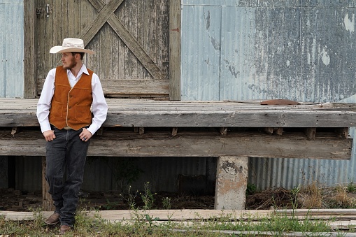 Cowboy, dressed in boots, hat, leans, and vest, confidently stands in front of a rustic barn door in Texas. Copy space.