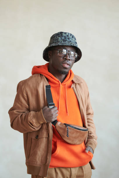 Medium studio portrait shot of handsome young Black man wearing stylish casual hoodie, jacket, bucket hat and belt bag looking at camera Stylish Black Man Studio Portrait bucket hat stock pictures, royalty-free photos & images