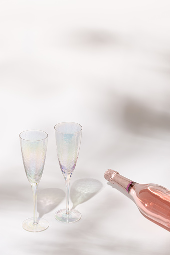 Ð¡losed bottle of rose champagne sparkling wine and empty glasses with sunlight on light table, celebration or relaxing evening for two people at summer day.