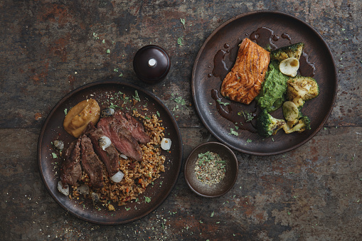 Grilled tenderloin bull steak with spelt grain,vegetables and smoked onion puree. Grilled salmon with broccoli-pea puree and steamed broccoli.  Flat lay top-down composition on dark background.