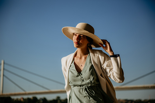 Beautiful portrait of a young woman standing, wearing a hat and looking at the Sun, radiating joy with her soft smile on face