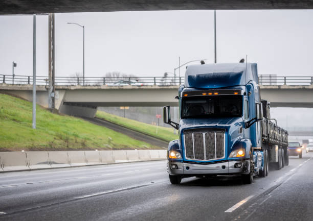 istock Blue classic industrial big rig semi truck transporting covered cargo on flat bed semi trailer running on the wide highway road at wet rain weather 1310853707