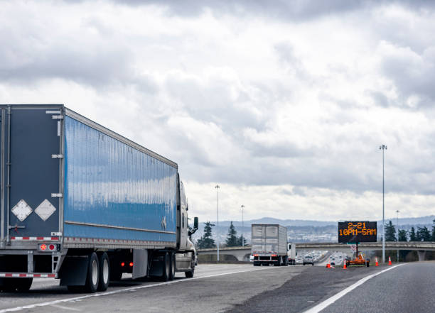 istock Convoy of big rigs semi trucks with different semi trailers running on the interstate highway with bridge and road sign electronic pane 1310854747