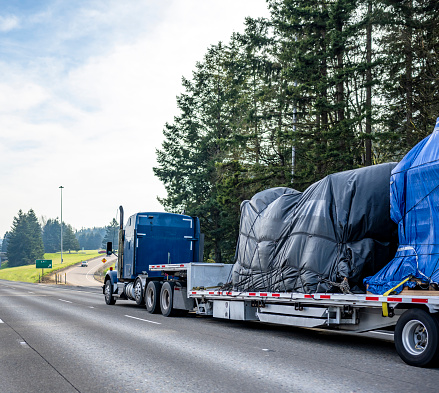 Huge blue classic big rig industrial semi truck transporting covered with tarp heavy commercial cargo on step down semi trailer running on the wide highway road with trees on the side