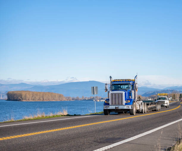 istock Blue powerful big rig semi truck with step down semi truck for oversize load transportation and escort car with oversize load sign on the roof driving on the road along the river 1310854857