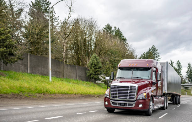 istock Burgundy low cab big rig semi truck transporting cargo in low profile covered semi trailer running on the multiline highway road with hill  and fence on the side 1310854945