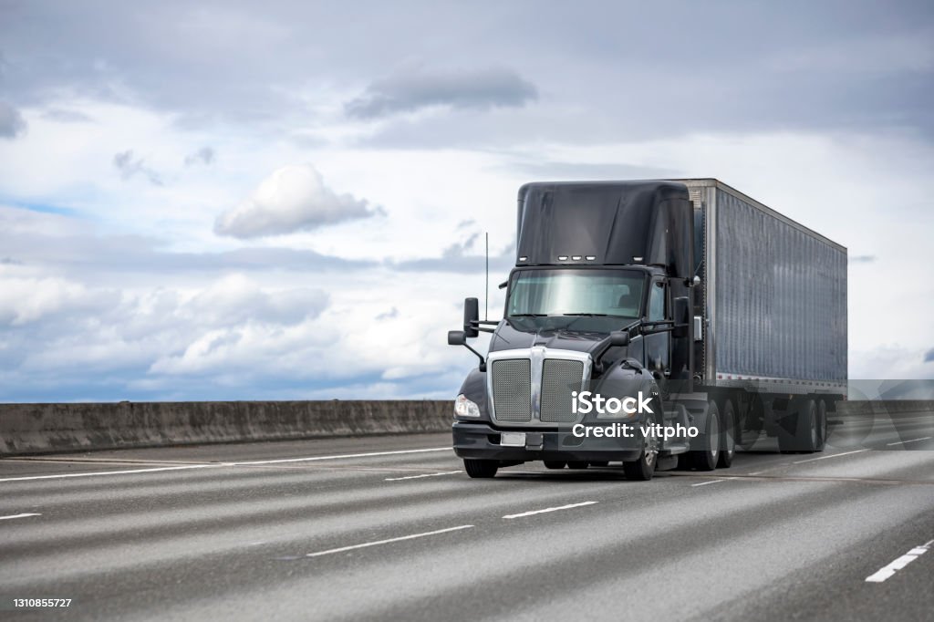 Powerful stylish black big rig day cab semi truck deliver commercial cargo in covered dry van semi trailer driving on the interstate highway road Commercial big rig black industrial grade day cab professional semi truck transporting cargo in dry van semi trailer driving on the interstate highway road with one way traffic direction Semi-Truck Stock Photo