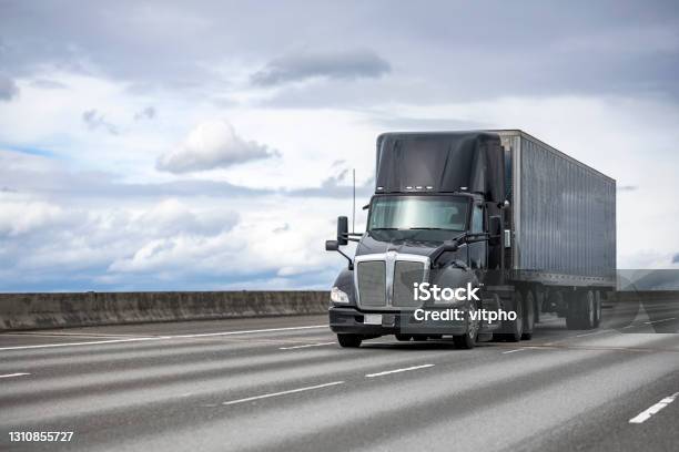 Powerful Stylish Black Big Rig Day Cab Semi Truck Deliver Commercial Cargo In Covered Dry Van Semi Trailer Driving On The Interstate Highway Road Stock Photo - Download Image Now
