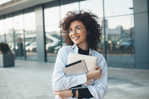 istock Curly haired business lady is embracing and holding a laptop while posing with glasses in front of a building 1310865892