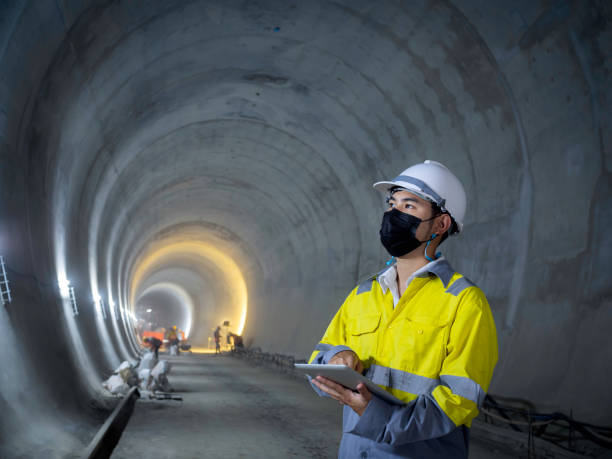 Young Asian tunnel engineering working at construction site. Young Asian tunnel engineering wearing high visibility jacket, face mask and white safety helmet working and using digital tablet in dark railway tunnel construction site area with copy space. tunnel stock pictures, royalty-free photos & images