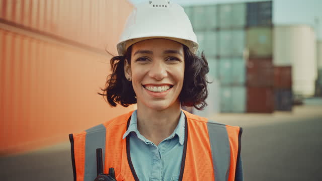 Smiling Portrait of a Beautiful Hispanic Female Industrial Engineer in White Hard Hat, Safety Vest and with Two-Way Radio Working in Logistics Center. Inspector or Supervisor in Container Terminal.