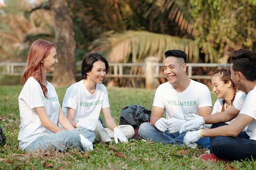 Group of smiling young men and women sitting on grass in city park and discussing volunteering