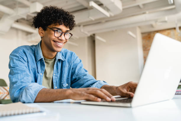 gelukkige glimlachende afro zakenman die laptop met behulp van bij het bureau in bureau - male employee office stockfoto's en -beelden