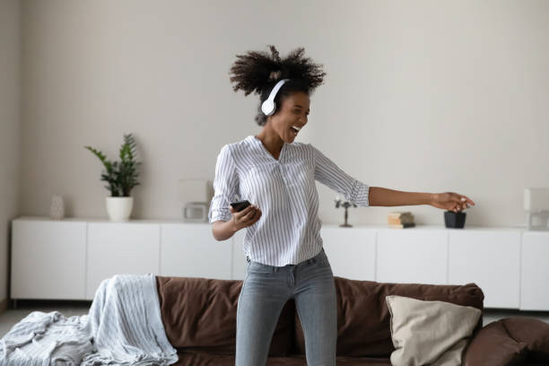 Excited biracial woman in headphones listen to music on cell Excited young African American woman in modern headphones listen to music on smartphone dance at home. Happy biracial female in earphones have fun enjoy good quality sound on cellphone gadget. roots music stock pictures, royalty-free photos & images