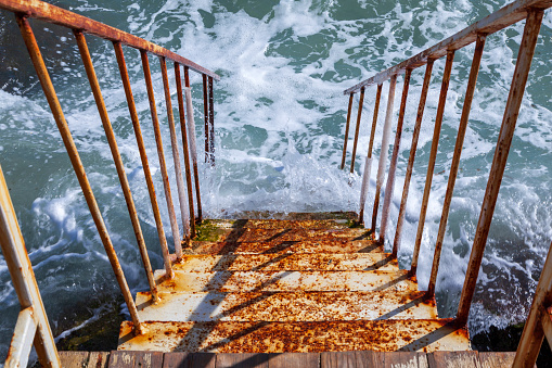 A rusty staircase goes into the blue water of the sea. Descent.