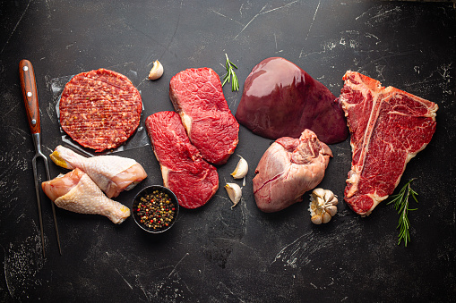 Selection of assorted raw meat food with seasonings for zero carb carnivore diet: uncooked beef steak, ground meat patty, heart, liver and chicken legs on black stone background from above