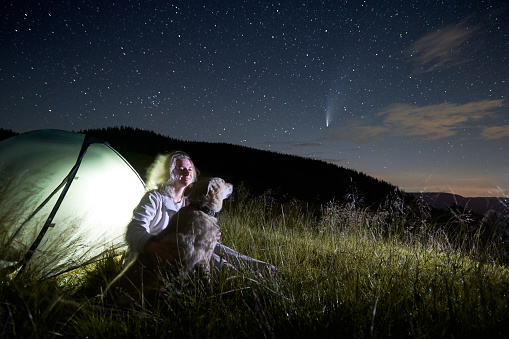 Young woman in the mountains observing beautiful starry night and a comet, sitting with her dog next to illuminated tent at campsite. Copy space. Concept of astrophotography