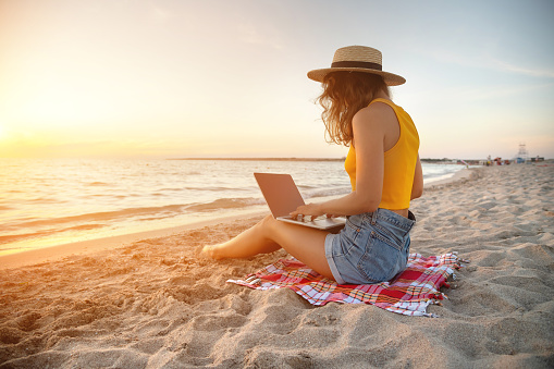 A slender young lady in denim shorts, a yellow tank top and a straw hat sits on a blanket on the sandy seashore and works on her laptop at sunset. Freelance and remote work and vacation concept.