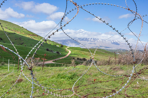 View of Mount Hermon with a snow-capped peak in the clouds through barbed wire. Israel