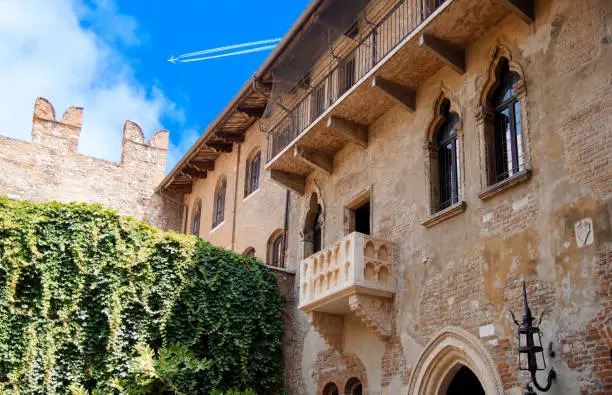 Photo of Romeo and Juliet balcony. Courtyard of Casa di Giulietta (House of Juliet or House of Cappelletti) against with Jet plane, Airliner in high flight with vapor trail.