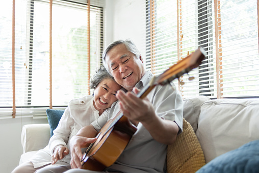 Happy Asian senior Couple enjoying singing and playing acoustic guitar together on sofa at home. Joyful Grandfather and Grandmother celebrating their Wedding Anniversary.