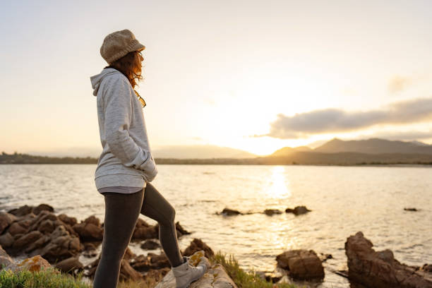 da sola donna pensierosa in piedi sulle rocce marine guardando al tramonto o all'alba tra la montagna all'orizzonte. casual bella persona con cappello di lana trova la sua spiritualità vivendo la natura in vacanza di viaggio - destinies foto e immagini stock