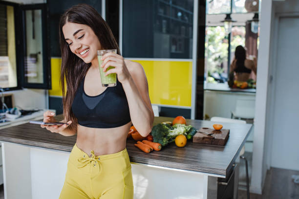 joven atlética con ropa deportiva bebe una bebida saludable en casa después de entrenar y usar el teléfono móvil - healthy eating sport exercising women fotografías e imágenes de stock