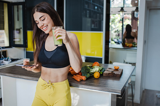 Athletic young woman in sportswear makes a healthy drink at home and enjoying after training while drinking a smoothie while using mobile phone
