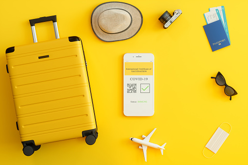 Digital Covid-19 Vaccine Passport App On Mobile Phone Screen. Passport, Airplane Tickets, Yellow Suitcase, And Protective Face Mask With On Yellow Background.