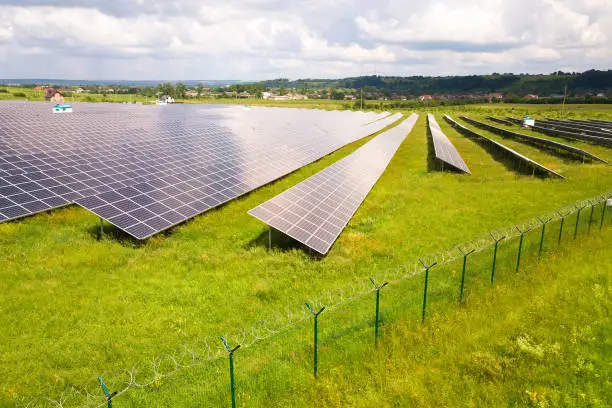 Photo of Aerial view of solar power plant on green field with protective wire fence around it. Electric panels for producing clean ecologic energy.