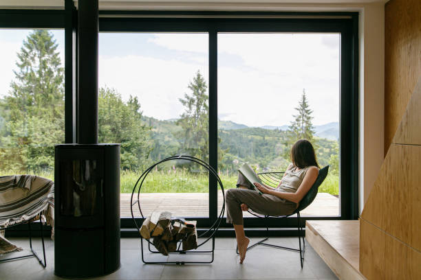 Beautiful stylish woman reading book, sitting on chair at fireplace on background of mountain hills Beautiful stylish woman reading book on chair at fireplace with firewood on background of mountain hills. Young female in casual clothes relaxing in modern chalet with amazing view from window chalet stock pictures, royalty-free photos & images