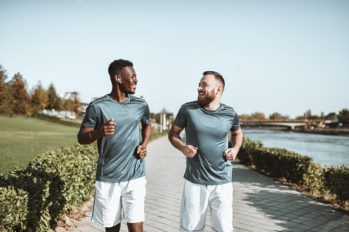 Two Smiling Males Of Different Ethnicities Running Together Near Park And River Quay