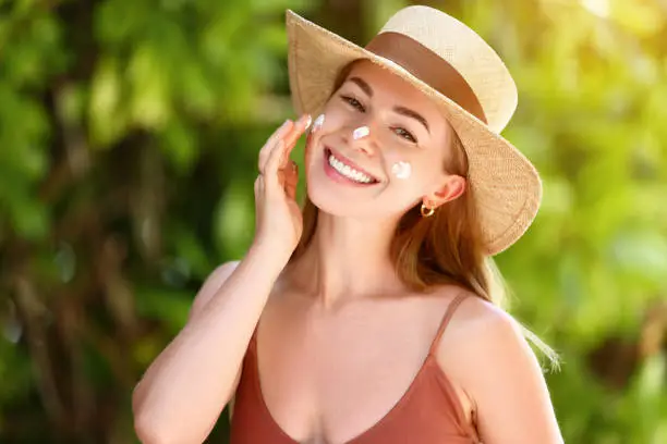 Smiling female wearing swimsuit with straw hat and smearing sunblock lotion on face looking at camera on blurred background
